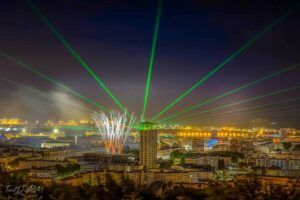 Europe Évènement - Photo de la tour d'une ville avec projection de faisceaux lasers verts partant de la tour et feux d'artifices au Havre